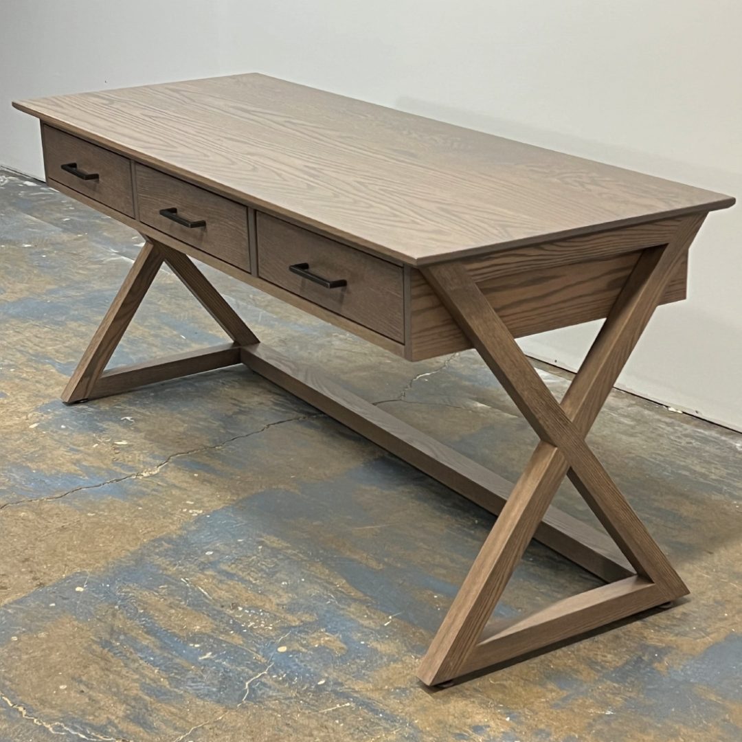 A Simple Guide To Wood Types For Furniture 4 A Modern Writing Desk With A Grey-Brown Wood Finish, Viewed From The Side. The Desk Stands On Unique X-Shaped Wooden Legs And Features A Flat Tabletop With Three Slender Drawers Across The Front For Storage.
